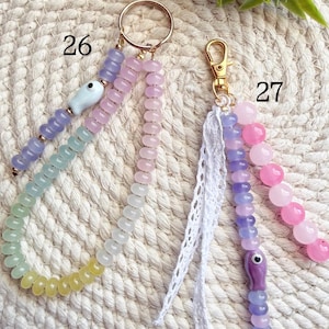 May include: Two beaded keychains. One has a gold ring and a white ceramic fish charm. The other has a gold clasp and a white lace tassel. Both have colorful beads in pastel shades of pink, purple, green, and yellow.