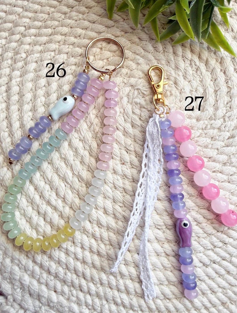 May include: Two beaded keychains. One has a gold ring and a white ceramic fish charm. The other has a gold clasp and a white lace tassel. Both have colorful beads in pastel shades of pink, purple, green, and yellow.
