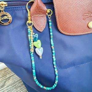 May include: A navy blue handbag with a brown leather flap and handle. A gold-toned keychain with turquoise and gold beads and three heart-shaped charms hangs from the handle. The keychain is attached with a gold clasp.