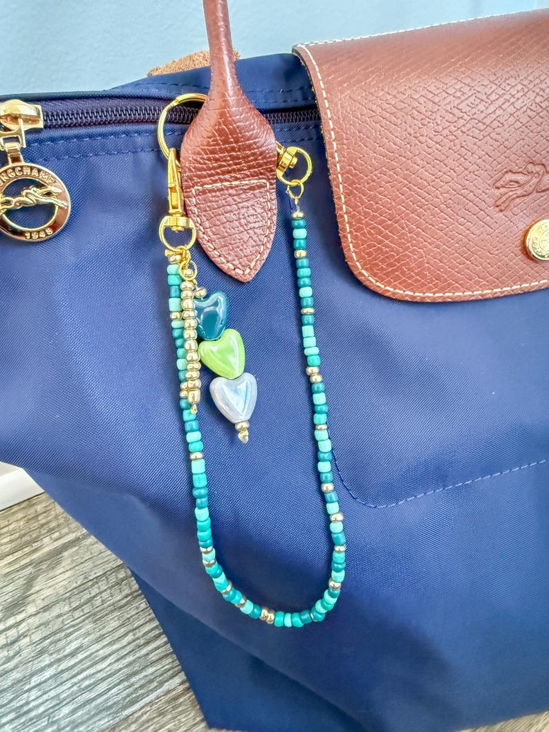 May include: A navy blue handbag with a brown leather flap and handle. A gold-toned keychain with turquoise and gold beads and three heart-shaped charms hangs from the handle. The keychain is attached with a gold clasp.