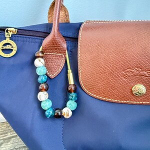 May include: A navy blue tote bag with a brown leather handle and flap. A beaded charm with brown, white, and turquoise beads hangs from the handle. A gold-colored zipper and emblem are visible.