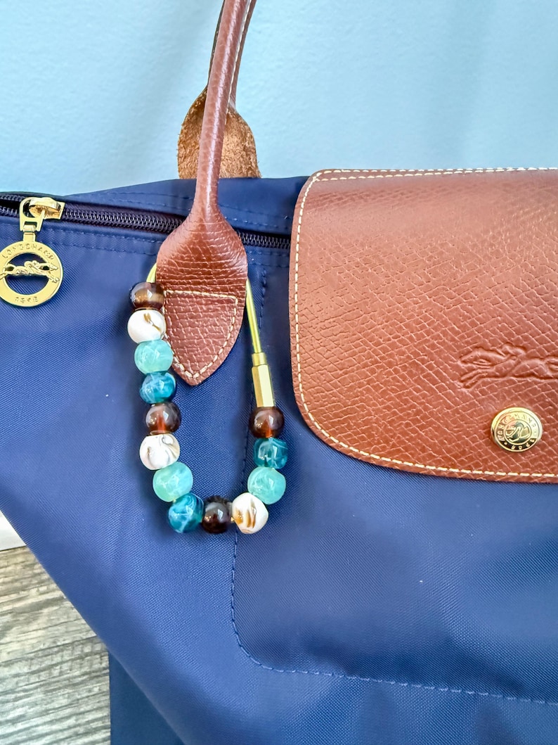 May include: A navy blue tote bag with a brown leather handle and flap. A beaded charm with brown, white, and turquoise beads hangs from the handle. A gold-colored zipper and emblem are visible.