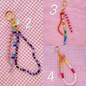 May include: Four beaded keychains with gold-colored hardware. Keychains feature various colors of beads, including pink, purple, blue, orange, and yellow. One keychain has a star charm. The keychains are on a pink and white checkered background.