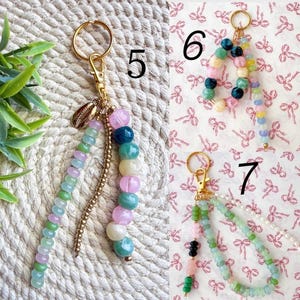 May include: Keychains with colorful beaded accents. One keychain features a gold-colored clasp, a small shell charm, and a strand of green, purple, and pink beads. Another has a strand of multi-colored beads. A third keychain has green and pink beads.