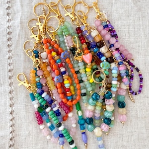 May include: A collection of beaded keychains in various colors, including orange, green, pink, and blue. Each keychain features a gold-colored clasp and a unique arrangement of beads, some with heart-shaped charms.