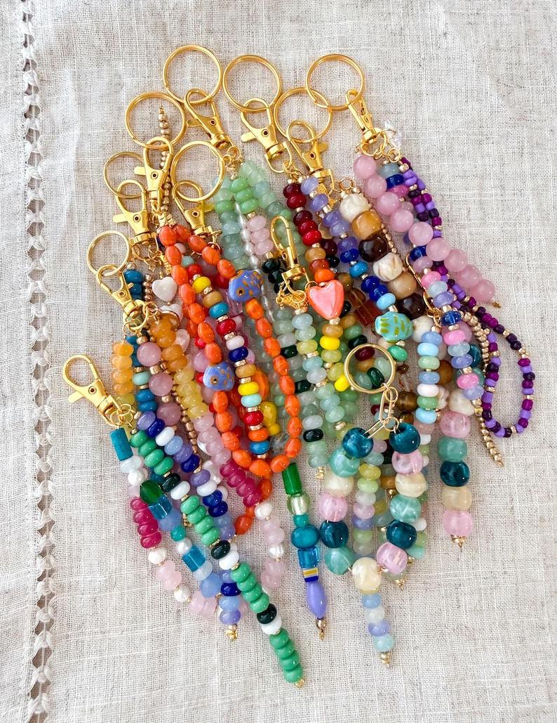 May include: A collection of beaded keychains in various colors, including orange, green, pink, and blue. Each keychain features a gold-colored clasp and a unique arrangement of beads, some with heart-shaped charms.