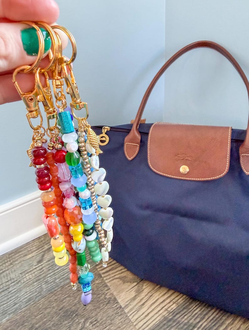 May include: A collection of colorful beaded keychains with gold-tone hardware. The keychains feature a variety of beads in rainbow hues, including red, orange, yellow, green, blue, and purple. A navy blue tote bag with brown leather accents is in the background.