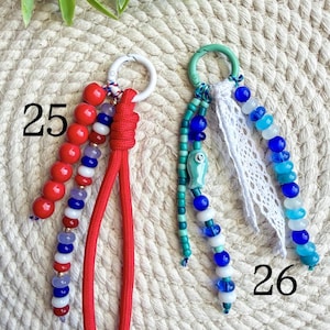 May include: Two keychains with beaded accents. One features red beads and a red cord, the other has blue and white beads with a fish charm and white lace. Numbers 25 and 26 are visible.