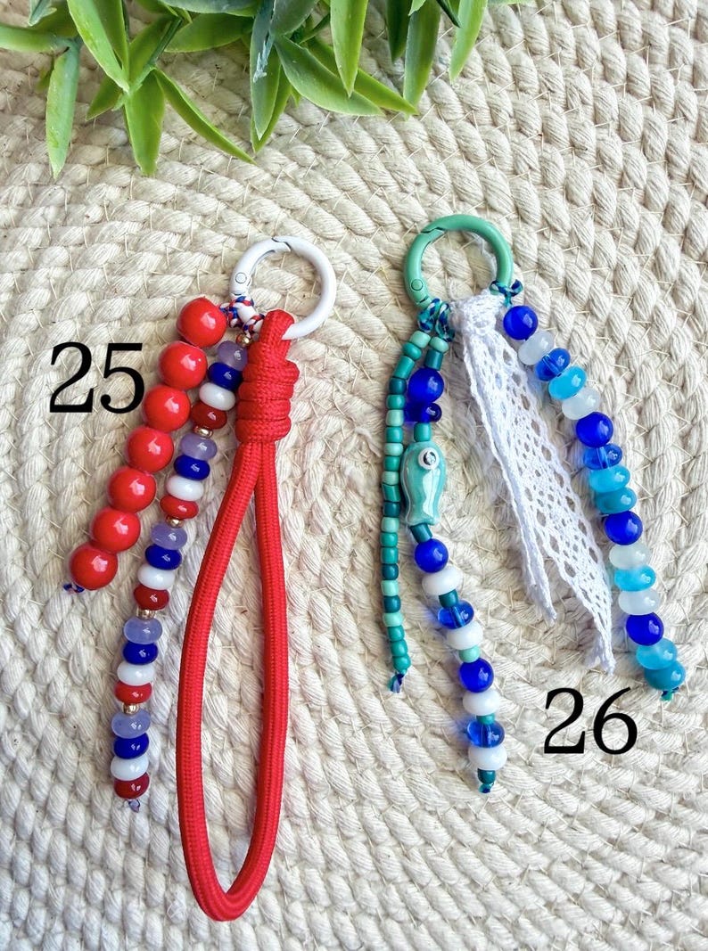 May include: Two keychains with beaded accents. One features red beads and a red cord, the other has blue and white beads with a fish charm and white lace. Numbers 25 and 26 are visible.