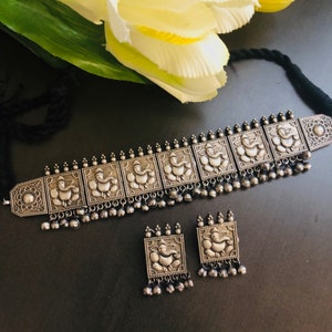 May include: A silver choker necklace and earrings set featuring an ornate design of the Hindu deity Ganesha. The necklace has a row of square-shaped pendants with Ganesha figures, and the earrings are square-shaped with a Ganesha figure in the centre.