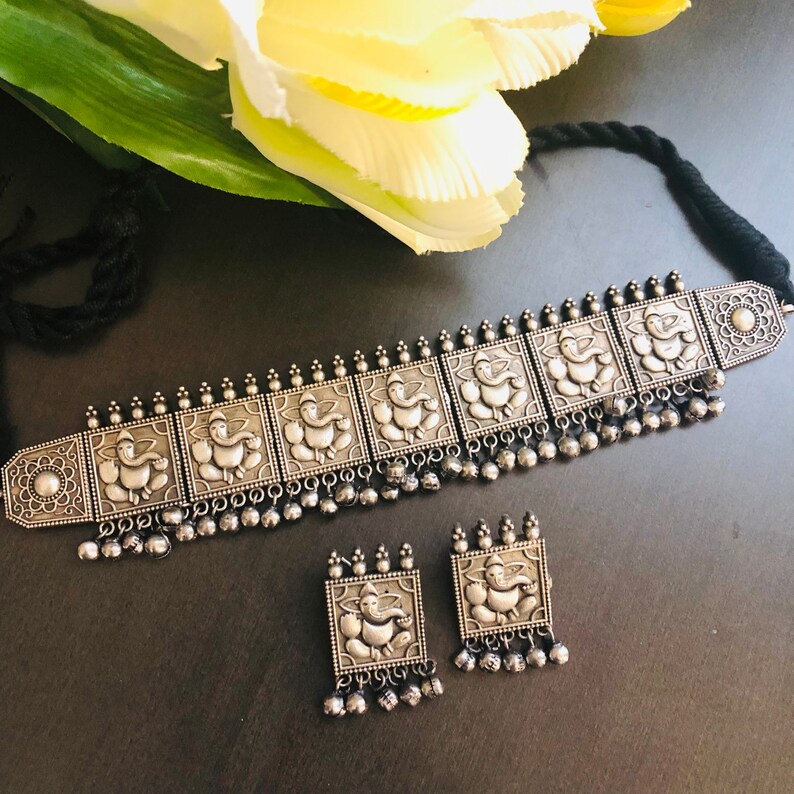 May include: A silver choker necklace and earrings set featuring an ornate design of multiple square-shaped pendants with an embossed image of the Hindu deity Ganesha. The necklace has a black string closure and the earrings have small silver bells hanging from them.