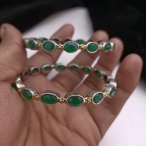 May include: Two silver bangles with green oval stones and gold accents. The bangles are stacked on top of each other.