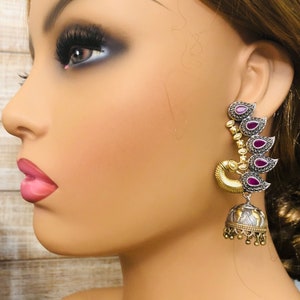 Pode incluir: Um par de brincos de prata e ouro com pedras roxas. Os brincos t&ecirc;m a forma de uma pena de pav&atilde;o e t&ecirc;m um pingente em forma de sino.