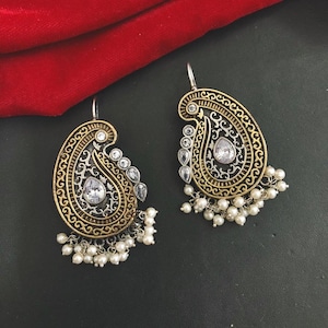 Puede incluir: Un par de pendientes de paisley en oro y plata con detalles de perlas blancas. Los pendientes tienen un diseño en forma de lágrima con detalles intrincados.