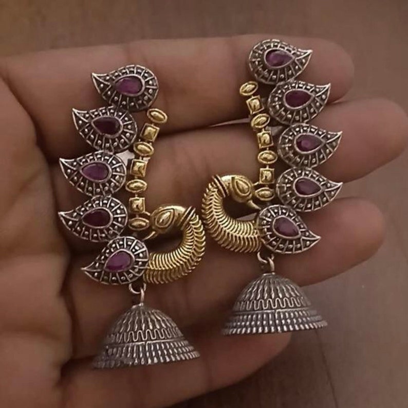 Pode incluir: Um par de brincos de prata e ouro com um pingente de sino. Os brincos apresentam um design de m&uacute;ltiplos pingentes em forma de l&aacute;grima com pedras vermelhas e um design de pav&atilde;o.