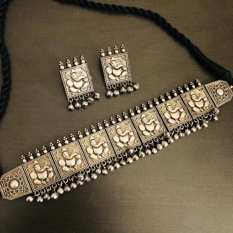 May include: A silver choker necklace with a black cord and silver earrings. The necklace features a repeating pattern of square-shaped pendants with an embossed image of the Hindu deity Ganesha. The earrings are also square-shaped with an embossed image of Ganesha.
