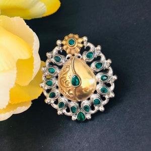 May include: A silver and gold ring with a teardrop-shaped design. The ring is decorated with green gemstones and intricate filigree work.