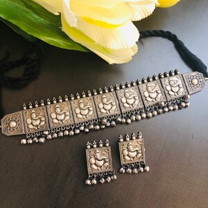 May include: A silver-toned choker necklace with a row of square pendants featuring an embossed design of the Hindu deity Ganesha. The necklace is accented with small silver beads and comes with a matching pair of earrings.
