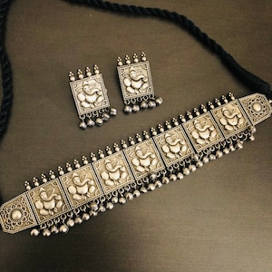 May include: A silver choker necklace with a black cord and silver earrings. The necklace features a repeating pattern of square-shaped pendants with an embossed image of the Hindu deity Ganesha. The earrings are also square-shaped with an embossed image of Ganesha.