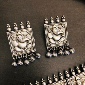 May include: A pair of silver earrings with a square design featuring an embossed image of the Hindu deity Ganesha. Each earring has a dangling row of small silver bells.
