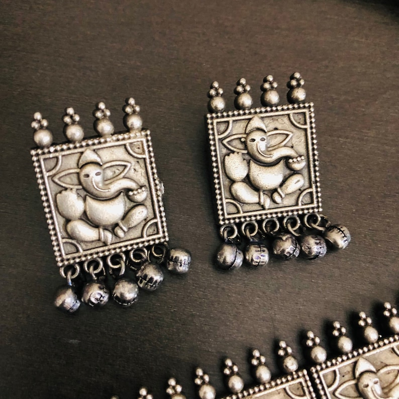 May include: A pair of silver earrings with a square design featuring an embossed image of the Hindu deity Ganesha. Each earring has a dangling row of small silver bells.