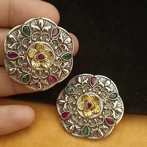 May include: A pair of silver earrings with intricate floral designs and colourful gemstones. The earrings feature a central flower with a red gemstone surrounded by smaller green and red gemstones.