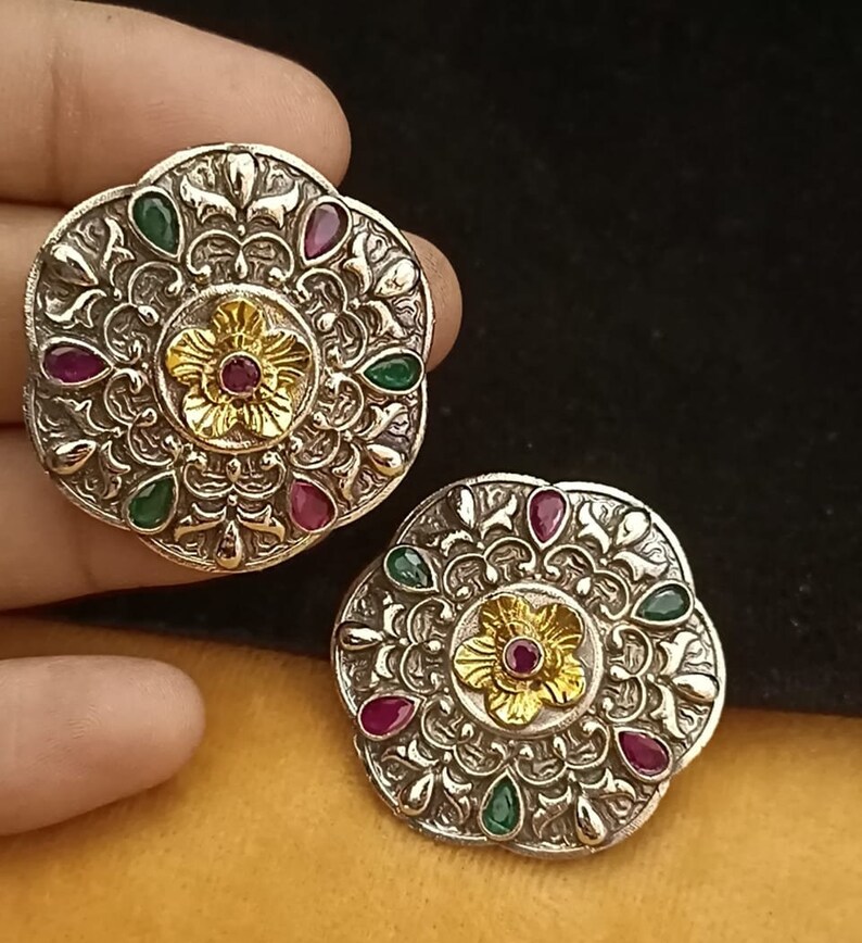 May include: A pair of silver earrings with intricate floral designs and colourful gemstones. The earrings feature a central flower with a red gemstone surrounded by smaller green and red gemstones.