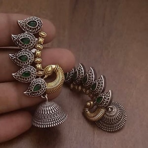 Pode incluir: Um par de brincos de prata e ouro com pedras verdes. Os brincos t&ecirc;m a forma de pav&atilde;o com as caudas abertas. Os brincos t&ecirc;m um sino pendurado na parte inferior.