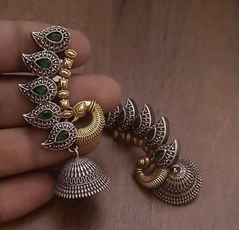 Pode incluir: Um par de brincos de prata e ouro com pedras verdes. Os brincos t&ecirc;m a forma de pav&atilde;o com as caudas abertas. Os brincos t&ecirc;m um sino pendurado na parte inferior.