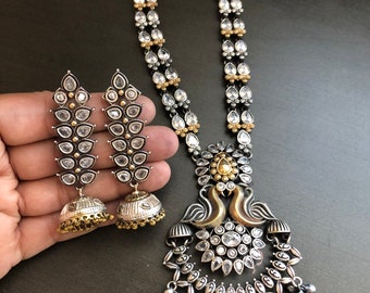 May include: A silver and gold necklace with a peacock design pendant and matching earrings. The necklace features a double strand of clear stones and the pendant has a floral design with a gold center. The earrings are long and feature a similar design to the pendant.