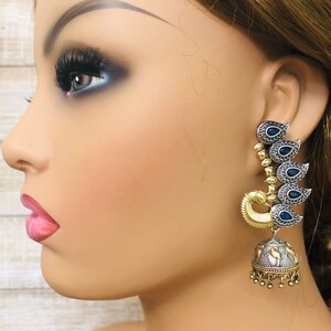 Pode incluir: Brincos pendentes em tom prata e dourado com detalhes de pedras preciosas azuis. Os brincos t&ecirc;m a forma de uma l&aacute;grima com um charme em forma de sino na parte inferior.