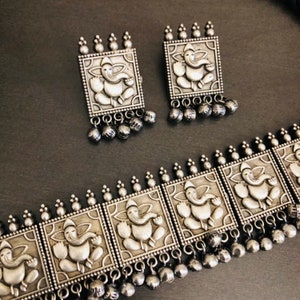 May include: Silver-toned necklace and earrings set featuring square pendants with an embossed image of the Hindu deity Ganesha. The necklace has multiple pendants and dangling beads.