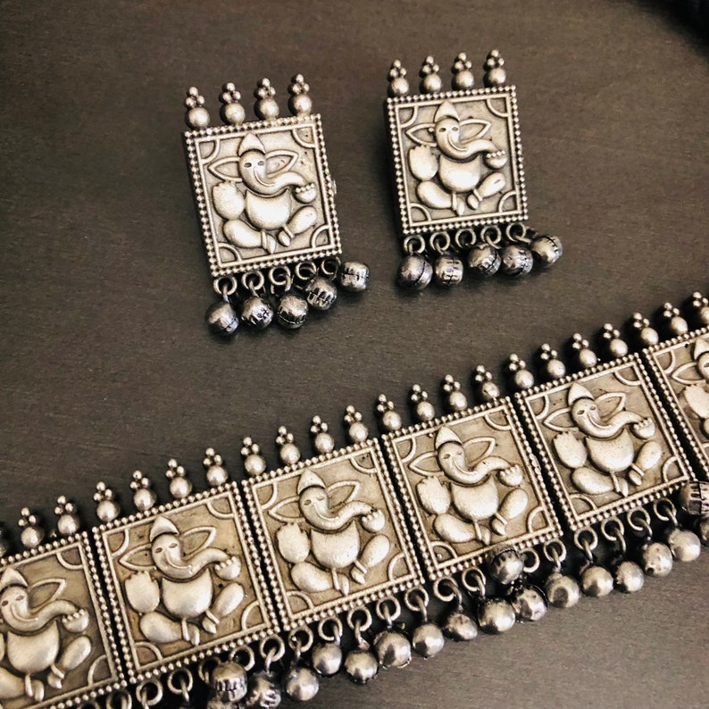 May include: Silver-toned necklace and earrings set featuring square pendants with an embossed image of the Hindu deity Ganesha. The necklace has multiple pendants and dangling beads.