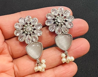 May include: A pair of silver earrings with a white flower design and a white teardrop-shaped stone hanging from each. The earrings have a cluster of white pearls hanging from the bottom.