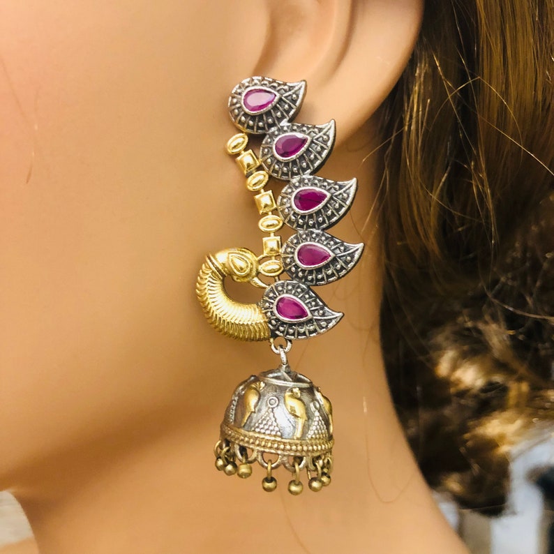 Pode incluir: Brincos de tom prateado e dourado com pedras rosa e design de pav&atilde;o. Os brincos t&ecirc;m um charme de sino pendurado.