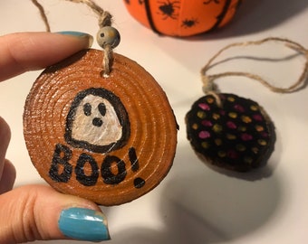 May include: Two wooden ornaments with painted Halloween designs. One ornament is brown with a white ghost and the text "BOO!" in black. The other ornament is black with pink and yellow polka dots.