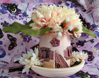 May include: A small, pink, house-shaped planter with a brown door and windows. The planter is filled with a bouquet of cream-colored flowers with green leaves.