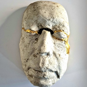 May include: A weathered, off-white mask with a human face. The mask has a crack across the forehead and eye area, filled with gold leaf. The mask has a rough, textured surface and is likely made of plaster or a similar material.