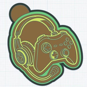 May include: A brown and light green gaming-themed cookie cutter featuring a headset with an attached microphone and a video game controller. The design is outlined in dark green and set against a light blue grid background with a circular top.