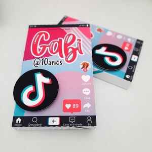 May include: Two decorative boxes with a social media theme. The boxes feature a pink, white, and blue color scheme with the name "Gabi" and the text "@10anos". Each box has a black circle with a white, red, and blue musical note logo.
