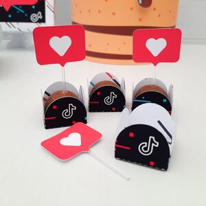 May include: A collection of decorated chocolates, each nestled in a black and white paper cup with a TikTok logo. Red heart-shaped toppers on clear sticks are placed above the chocolates. A large cake is visible in the background.