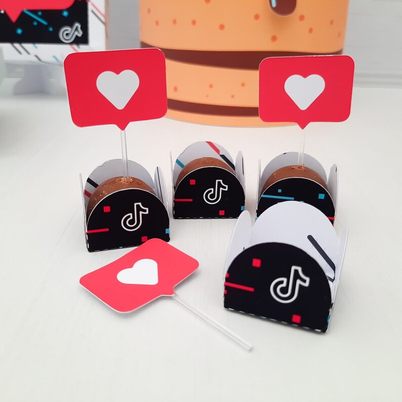 May include: A collection of decorated chocolates, each nestled in a black and white paper cup with a TikTok logo. Red heart-shaped toppers on clear sticks are placed above the chocolates. A large cake is visible in the background.