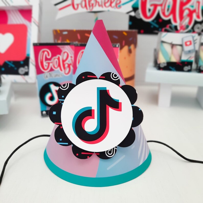 May include: A party hat with a TikTok logo. The cone-shaped hat features a color block design in pink, blue, and teal. A black string is attached to the base of the hat. The TikTok logo is a white circle with the app's logo in the center.