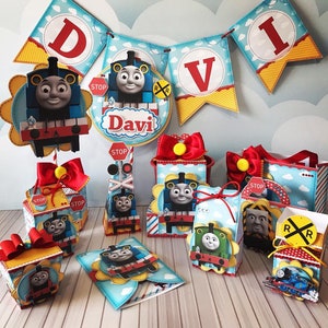 May include: A collection of Thomas the Tank Engine themed party decorations, including a banner spelling "DAVI", gift boxes, and a decorative centerpiece. The decorations feature the character Thomas, stop signs, and railroad crossing signs. The color scheme includes blue, red, yellow, and white.