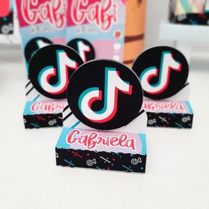 May include: A collection of party favors featuring the TikTok logo. The favors include round black and white stickers with the TikTok logo in red, white, and blue. The favors are displayed on small boxes with the name "Gabriela" in pink.