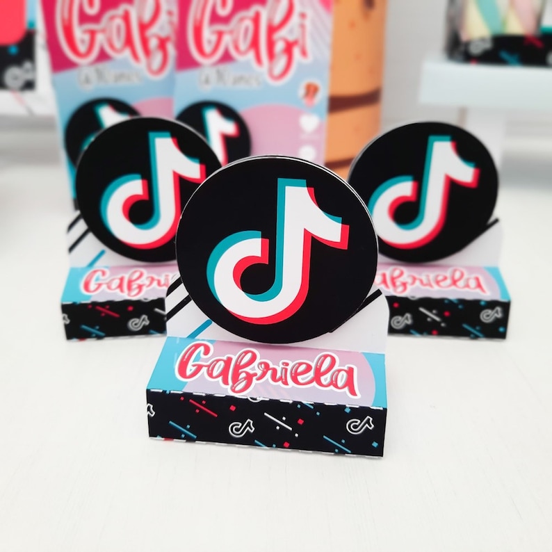 May include: A collection of party favors featuring the TikTok logo. The favors include round black and white stickers with the TikTok logo in red, white, and blue. The favors are displayed on small boxes with the name "Gabriela" in pink.