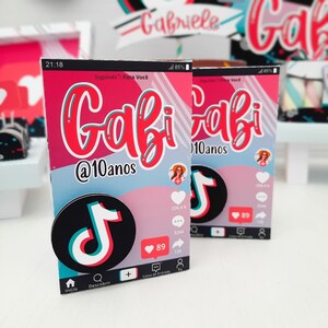 May include: Two party favor boxes with a pink and blue design, featuring the name "Gabi" in red script and the text "@10anos". Each box has a TikTok logo and social media icons. The boxes are for a birthday celebration.
