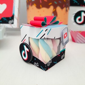 May include: A white, black, and blue box with a red bow on top, containing twisted marshmallows in pastel colors. The box has a clear window and a TikTok logo. A phone graphic with a heart is on the side. Other TikTok-themed decorations are in the background.