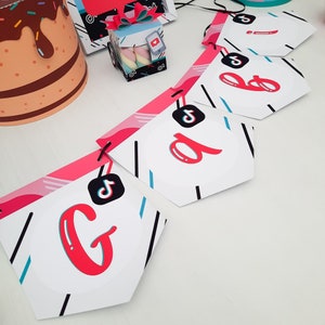 May include: A party banner with the letters "G B A" in red, white, and black. The banner has a TikTok logo on each pennant. Other party decorations include a cake-shaped hat and a gift box with marshmallows.