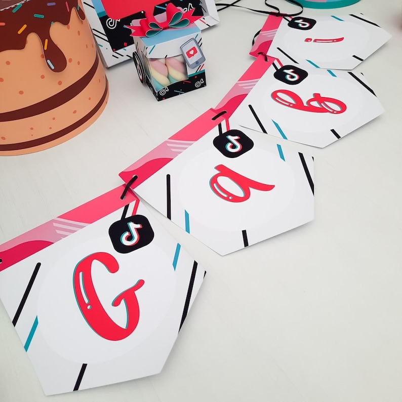 May include: A party banner with the letters "G B A" in red, white, and black. The banner has a TikTok logo on each pennant. Other party decorations include a cake-shaped hat and a gift box with marshmallows.